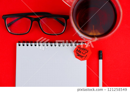A white notebook with a black spring at the top lies in the center of the photo, next to it is a white pencil with a black eraser, transparent black-framed glasses and a mug of tea are on top, a red 121258549