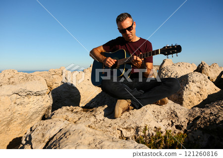 A man in a dark t-shirt playing acoustic guitar on a rocky area by the sea 121260816