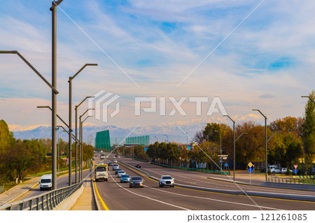A six-lane highway with entrances and exits against the backdrop of a picturesque sky and mountain ranges with snow caps in the background. Cars move in both directions, there are road signs 121261085