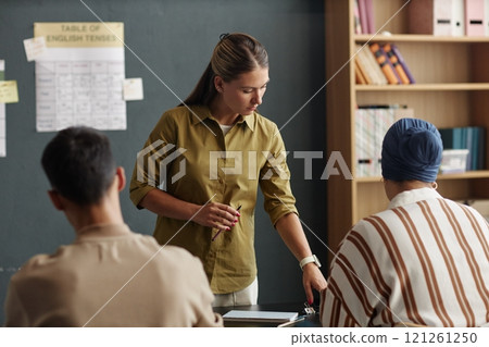 Young female English teacher conducting group class pointing at text in book drawing students attention to grammar rules at language school for adult learners, copy space 121261250