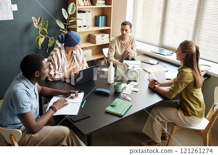 Adult man talking to English teacher and fellow students practicing new vocabulary during speaking club session while sitting at large desk in language school classroom 121261274