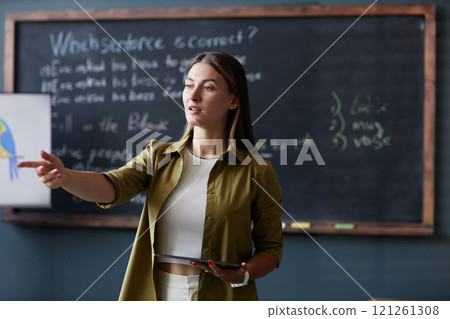 Medium shot of female English teacher holding tablet computer addressing student with question, while discussing new grammar rules in group class against blackboard at language school Medium shot of female English teacher holding tablet computer addressing student with question, while discussing new grammar rules in group class against blackboard at language school 121261308