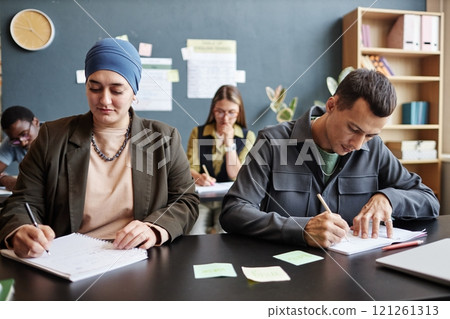 Medium shot of adult Muslim woman writing in exercise book sitting at desk with male fellow student writing test while studying English in language course group, copy space Medium shot of adult Muslim woman writing in exercise book sitting at desk with male fellow student writing test while studying English in language course group, copy space 121261313