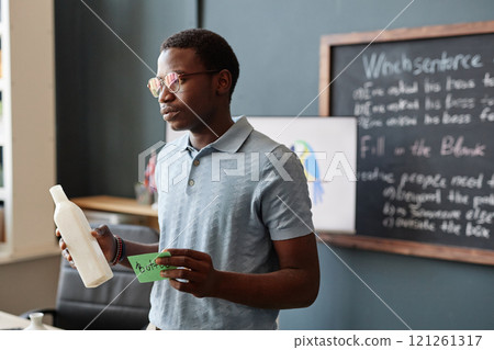 Medium shot of young African American teacher holding bottle and sticky note introducing new vocabulary to class using visual aids during lesson in English language school, copy space Medium shot of young African American teacher holding bottle and sticky note introducing new vocabulary to class using visual aids during lesson in English language school, copy space 121261317