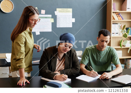 Medium shot of adult man and Muslum woman writing in notebooks sitting at desk with teacher supervising students during test in English language school, continuing education concept, copy space 121261338