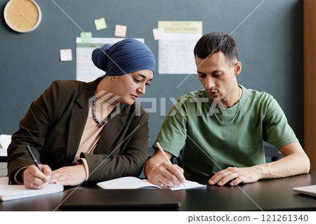 Medium shot of adult man explaining task to female Mulsim fellow student writing in exercise book while sitting at desk in English language school classroom Medium shot of adult man explaining task to female Mulsim fellow student writing in exercise book while sitting at desk in English language school classroom 121261340
