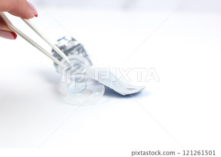 Soft blue contact lens lies on the edge of disposable contact lens storage container, next to it lies a sealed disposable lens storage container and a hand with tweezers reaches out to the lens to 121261501