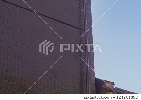 Wall of high-rise building without windows with dark chocolate light, visible texture of rows of brickwork against background of blue sky with haze. Pipe of roof drainage system is mounted at corner Wall of high-rise building without windows with dark chocolate light, visible texture of rows of brickwork against background of blue sky with haze. Pipe of roof drainage system is mounted at corner 121261868
