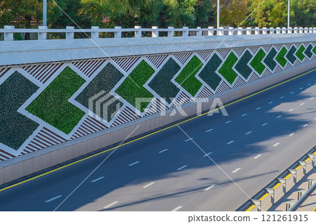The side direction signs on eight-lane highway are saddled with green artificial plants. On the asphalt there is discontinuous white and yellow solid strip, dividing metal bumpers, no cars, no people 121261915