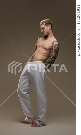 Full-length portrait of handsome young guy with shirtless, athletic, tattooed body design in grey sportive pants against neutral studio background Full-length portrait of handsome young guy with shirtless, athletic, tattooed body design in grey sportive pants against neutral studio background 121261951