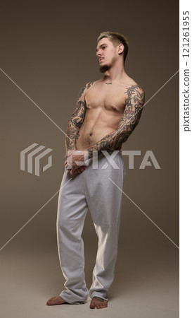 Full-length image of handsome young man with tattooed muscular body, posing shirtless in gray pants against beige studio background Full-length image of handsome young man with tattooed muscular body, posing shirtless in gray pants against beige studio background 121261955