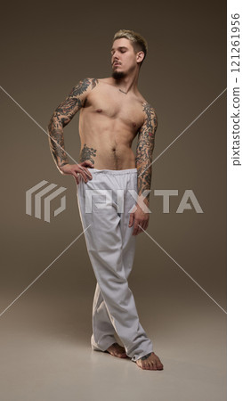 Full-length image of handsome young man with tattooed muscular body, posing shirtless in gray pants against beige studio background 121261956