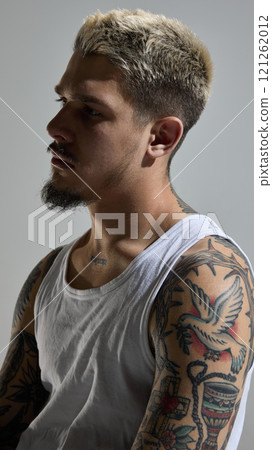 Portrait of handsome young man with blonde hair, beard and tattoos body, wearing tank top and posing with thoughtful expression Portrait of handsome young man with blonde hair, beard and tattoos body, wearing tank top and posing with thoughtful expression 121262012