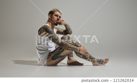 Side view of handsome shirtless young man with full-body tattoos, posing thoughtfully in white boxer briefs against neutral studio background. 121262014