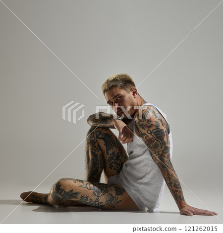 Tattooed male model sitting on floor wearing white tank top, showcasing detailed body tattoos in relaxed pose against neutral studio background Tattooed male model sitting on floor wearing white tank top, showcasing detailed body tattoos in relaxed pose against neutral studio background 121262015