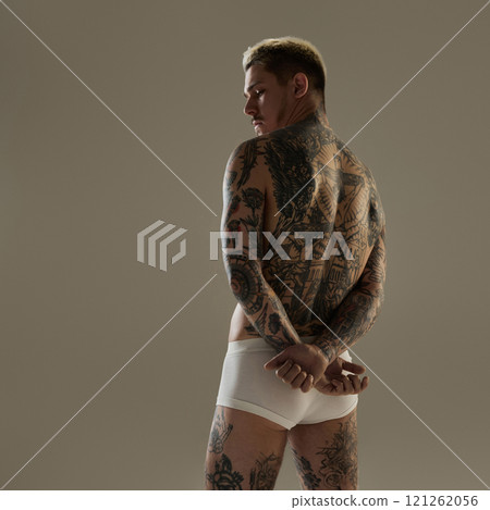 Rear view of tattooed male model with hands crossed behind his back, showing athletic tattooed body and strong physique in studio setting. 121262056