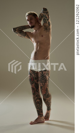 Full-length portrait of confident young young tattooed male model standing with his hands behind his head, emphasizing his full-body tattoos and strong physique Full-length portrait of confident young young tattooed male model standing with his hands behind his head, emphasizing his full-body tattoos and strong physique 121262062