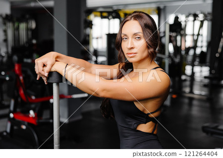 Female bodybuilder with long hair posing in gym with sports equipment. Strong woman with muscular body leaning on barbells in gym in dark atmosphere, looking at camera. 121264404