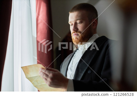 Side view portrait of classic gentleman in classic black suit holding letter while gazing at it near softly lit curtains, copy space Side view portrait of classic gentleman in classic black suit holding letter while gazing at it near softly lit curtains, copy space 121266528