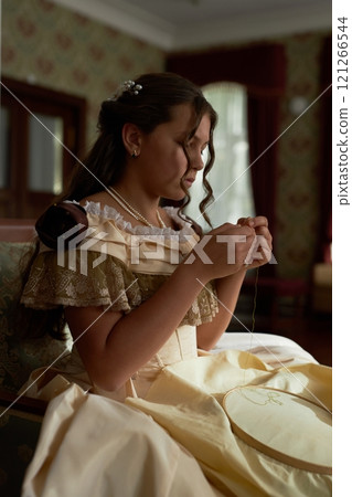 Vertical side view portrait of young girl wearing elegant dress with ruffles focusing on delicate embroidery in softly lit classic room Vertical side view portrait of young girl wearing elegant dress with ruffles focusing on delicate embroidery in softly lit classic room 121266544