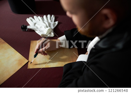 Over shoulder view of classic gentleman writing letter with ink on parchment over red velvet table, focus on hand holding fountain pen in soft natural light, copy space Over shoulder view of classic gentleman writing letter with ink on parchment over red velvet table, focus on hand holding fountain pen in soft natural light, copy space 121266548
