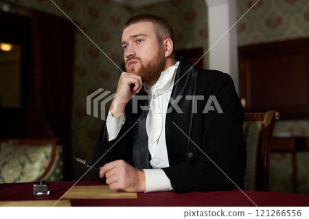 Portrait of young bearded gentleman wearing tailcoat looking away pensively while daydreaming at table in classic interior Portrait of young bearded gentleman wearing tailcoat looking away pensively while daydreaming at table in classic interior 121266556