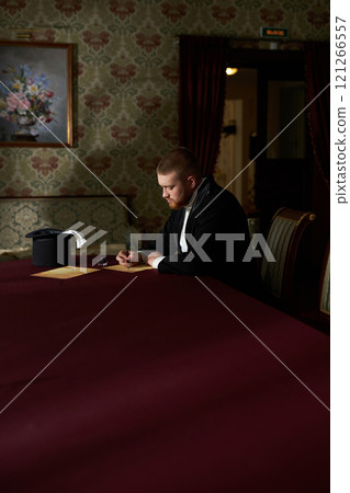 Vertical wide angle portrait of young man wearing tuxedo writing letter while sitting at table alone in classic interior with natural lighting Vertical wide angle portrait of young man wearing tuxedo writing letter while sitting at table alone in classic interior with natural lighting 121266557