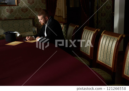Wide angle image of young gentleman writing letter with ink while sitting at red velvet table in classic interior, copy space Wide angle image of young gentleman writing letter with ink while sitting at red velvet table in classic interior, copy space 121266558