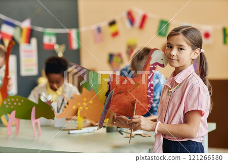 Side view portrait of cute young girl holding handmade dinosaur figure and smiling at camera in arts and crafts classroom copy space Side view portrait of cute young girl holding handmade dinosaur figure and smiling at camera in arts and crafts classroom copy space 121266580