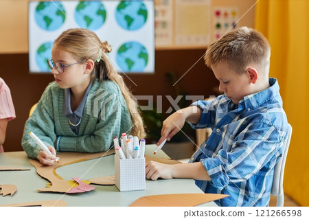 Side view portrait of blonde young boy painting handmade dinosaur figure while enjoying art and craft class with group of children in school copy space Side view portrait of blonde young boy painting handmade dinosaur figure while enjoying art and craft class with group of children in school copy space 121266598