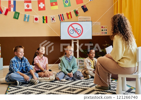 Wide angle scene of young teacher explaining road safety rules to cheerful group of children sitting on floor in colorful school classroom copy space Wide angle scene of young teacher explaining road safety rules to cheerful group of children sitting on floor in colorful school classroom copy space 121266740