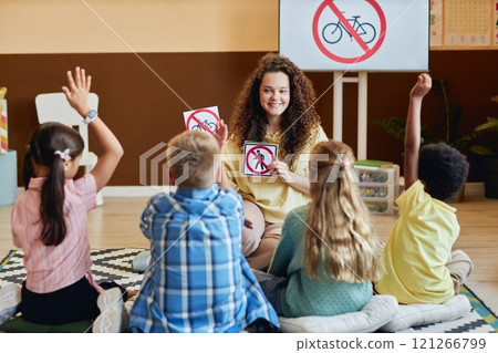 Front view of young female teacher holding road safety cards and smiling while teaching group of children in colorful primary school classroom 121266799