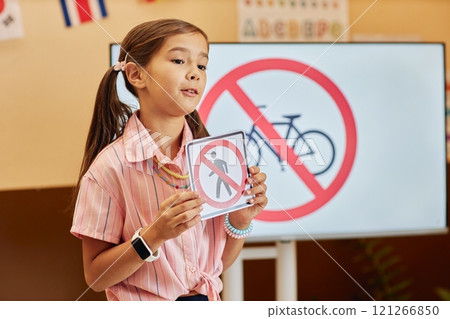 Waist up portrait of cute young girl with pigtails holding road safety sign while answering questions in primary school classroom copy space 121266850