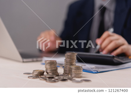 Stacks of coins with businessman calculating finances on desk in the background representing investment and saving 121267400