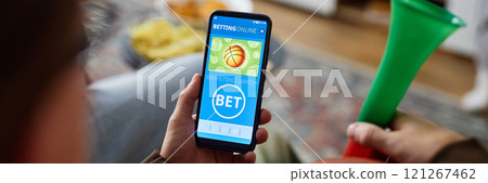 Header image of man holding smartphone with online sports bets app on screen while watching basketball game at home, copy space 121267462