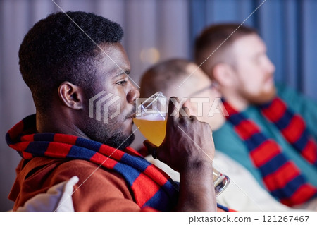 Side view closeup of adult African American man drinking beer from tall glass while sitting on couch and watching sports match on TV Side view closeup of adult African American man drinking beer from tall glass while sitting on couch and watching sports match on TV 121267467