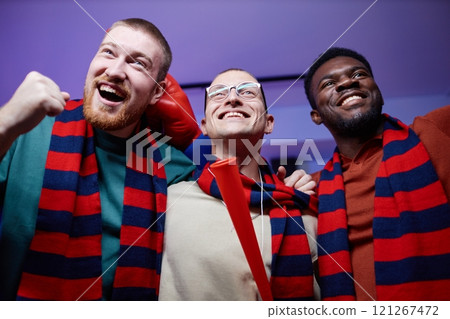 Low angle portrait of three excited sports fans huddling and cheering at home watching game match on TV and wearing matching scarves in team colors 121267472