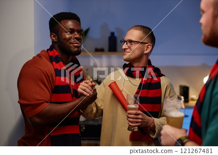 Front view portrait of three sports fans in team colored scarves smiling and shaking hands while celebrating victory in home kitchen setting Front view portrait of three sports fans in team colored scarves smiling and shaking hands while celebrating victory in home kitchen setting 121267481