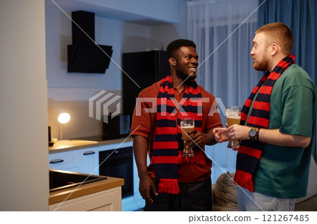 Side view portrait of two sports fans wearing matching team scarves talking and holding drinks discussing match in relaxed home setting, copy space 121267485