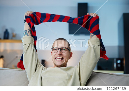Waist up portrait of excited sports fan cheering while holding striped scarf in team colors and watching match on TV at home, copy space Waist up portrait of excited sports fan cheering while holding striped scarf in team colors and watching match on TV at home, copy space 121267489