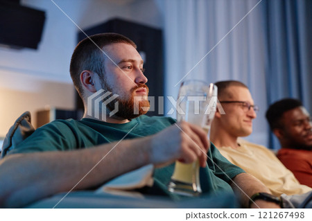 Side view portrait of bearded young man holding beer glass while watching TV with friends at home and holding beer glass, copy space Side view portrait of bearded young man holding beer glass while watching TV with friends at home and holding beer glass, copy space 121267498