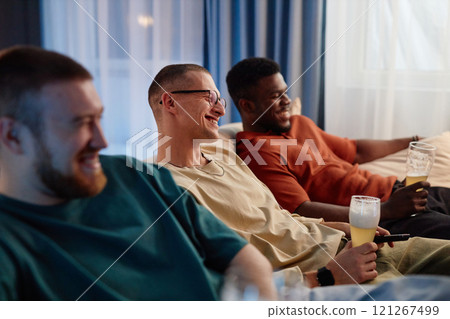 Side view portrait of three friends sitting on couch watching TV together and smiling holding beer glasses, copy space Side view portrait of three friends sitting on couch watching TV together and smiling holding beer glasses, copy space 121267499