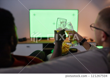 Back view of two men clinking beer glasses while sitting on couch and watching TV with blank chroma key green screen, copy space 121267504