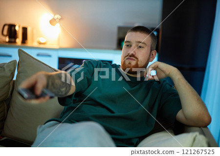 Front view portrait of relaxed bearded young man lounging on couch and holding TV remote in dimly lit living room, copy space Front view portrait of relaxed bearded young man lounging on couch and holding TV remote in dimly lit living room, copy space 121267525