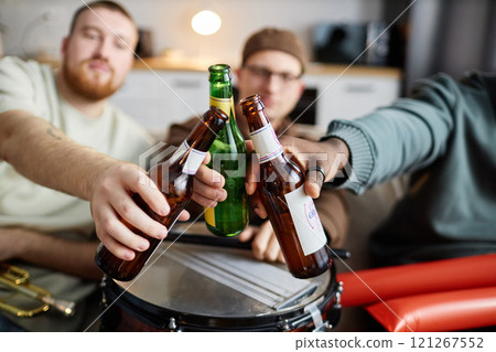 Close up of friends drinking beer with focus on hands clinking beer bottles in celebration while sitting on couch, copy space 121267552