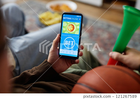 High angle close up of man holding smartphone with sports betting app on screen while watching basketball game on TV, copy space High angle close up of man holding smartphone with sports betting app on screen while watching basketball game on TV, copy space 121267589