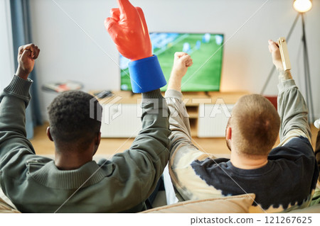 Back view of excited sports fans watching football match on TV at home sitting on couch and raising hands celebrating victory, copy space 121267625