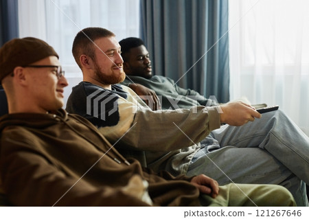 Side view portrait of smiling adult man watching TV with friends sitting on couch in row and switching channels using remote, copy space Side view portrait of smiling adult man watching TV with friends sitting on couch in row and switching channels using remote, copy space 121267646