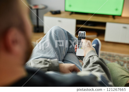 Over shoulder view of adult man watching TV at home with focus on hand holding remote, copy space 121267665