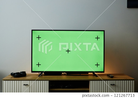 Front view background image of big flat screen TV with green chroma key screen on wooden Tv stand at home against white wall, copy space 121267713